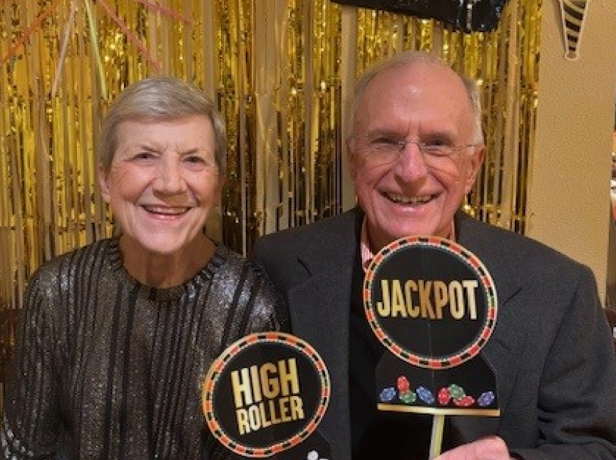 An older woman and man smile while holding signs that read HIGH ROLLER and JACKPOT in front of gold streamers and casino-themed decorations.