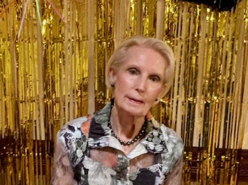 An older woman with short blonde hair, wearing a floral blouse and a black beaded necklace, stands in front of a gold tinsel curtain backdrop.