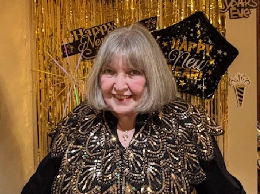 A smiling woman with gray hair wears a black and gold embellished top, standing in front of gold tinsel and Happy New Year decorations.