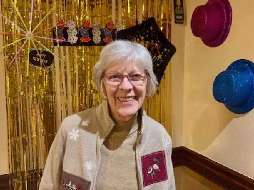 An older woman with gray hair and glasses smiles in front of gold tinsel and New Year’s decorations, including colorful hats on the wall and a festive Happy New Year sign.
