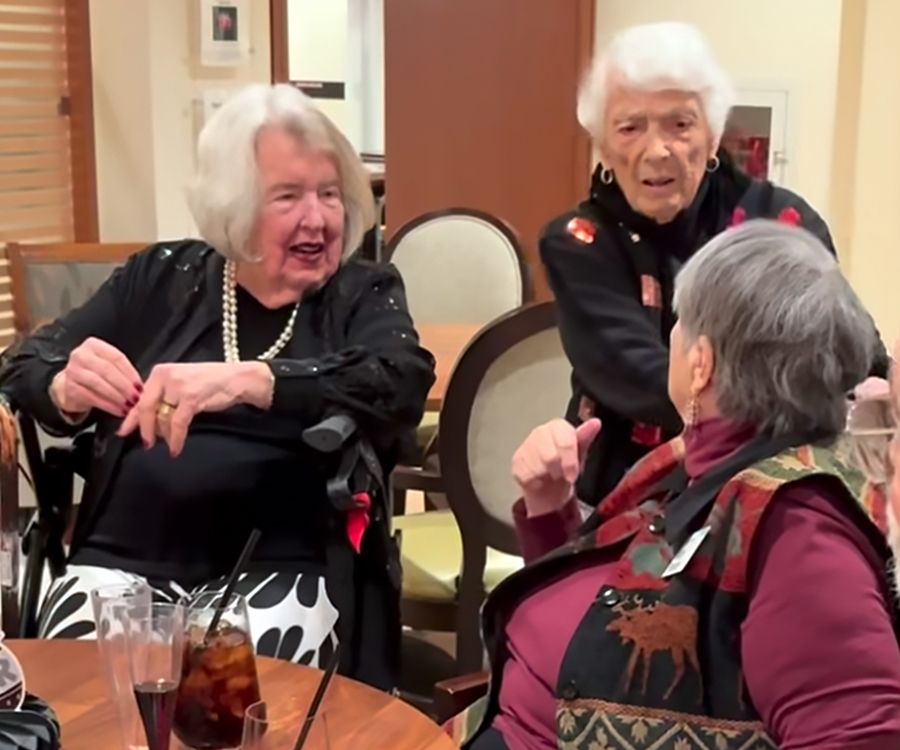 Three elderly women sit and chat around a table with drinks, appearing animated and cheerful. Two women face each other, smiling, while the third woman, standing, gestures as she talks to them.