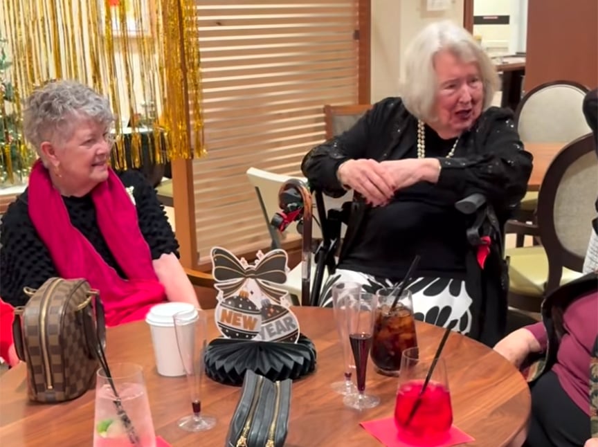 Two older women sit at a table decorated for New Year’s, with drinks and a Happy New Year sign. They are smiling and talking, dressed in festive clothing, with one woman sitting in a wheelchair.