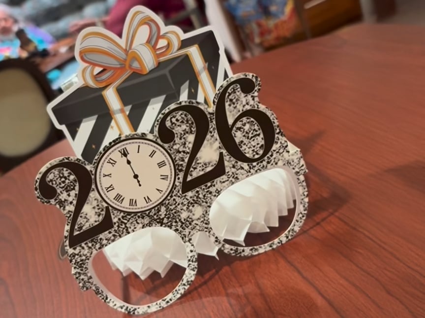 Novelty glasses with “2026” and a clock design sit on a wooden table, with a decorative black-and-white striped gift and bow on top. Blurred background shows a person and colorful objects.