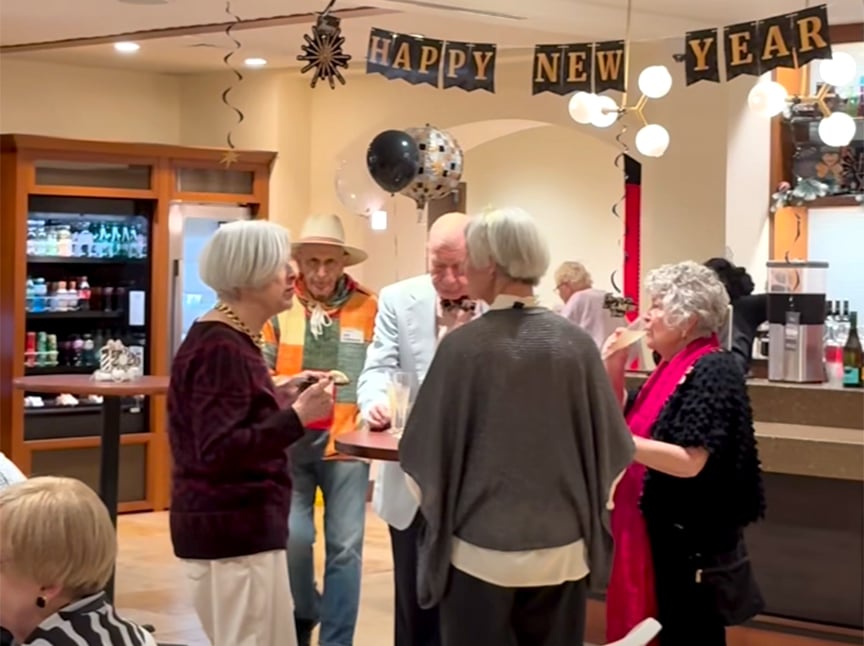 A group of older adults gather and chat at a New Year’s party. A Happy New Year banner hangs above, and festive decorations and balloons fill the room. Some people hold drinks while mingling near a counter.