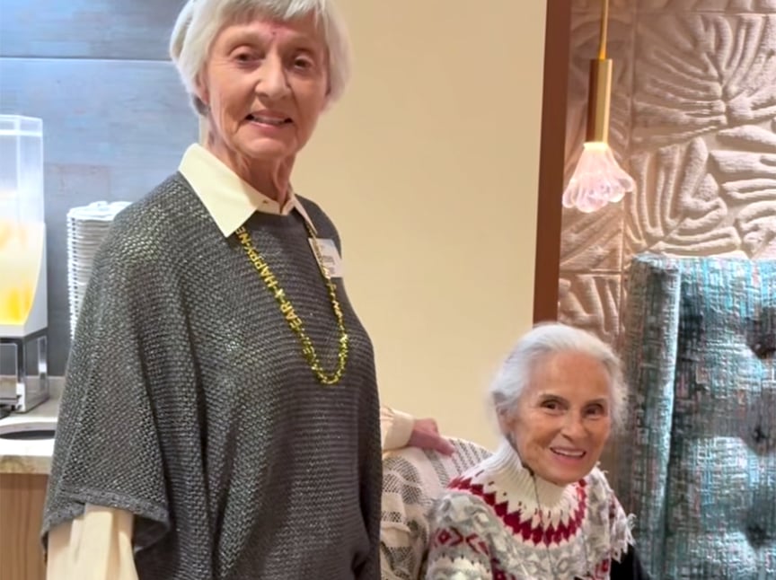 Two older women smiling indoors; one is standing and wearing a gray top with a gold necklace, while the other is seated and wearing a patterned sweater. The setting appears to be a cozy, warmly lit room.