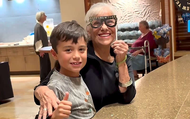 An older woman wearing “Happy New Year” glasses smiles and hugs a young boy who is giving a thumbs up. They are indoors, standing at a counter, with people and decorations in the background.