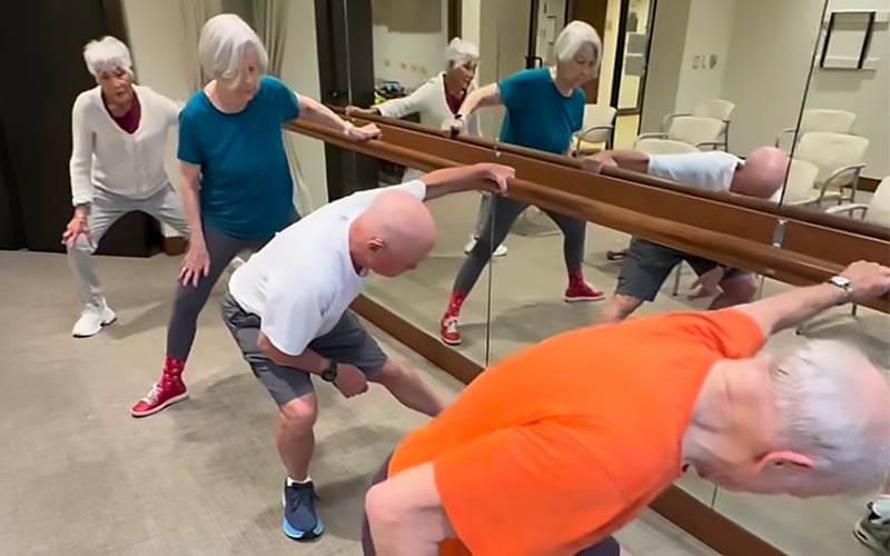 Four older adults exercise in a fitness room, stretching near a mirrored wall. Two women and two men are bending and reaching, dressed in casual athletic wear. Their reflections are visible in the mirror.