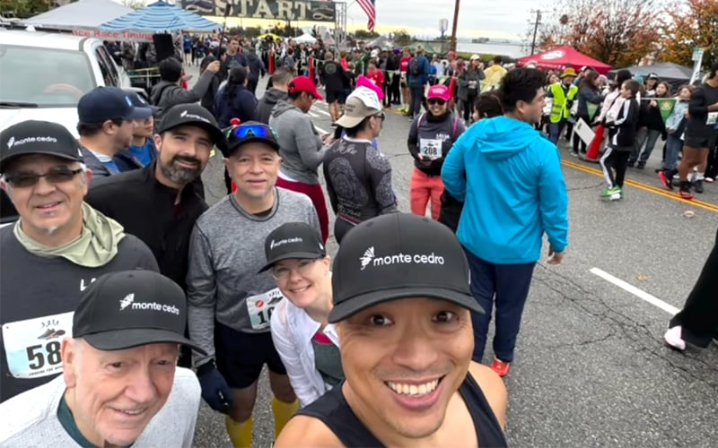 A group of smiling runners pose for a selfie at a crowded race starting line, all wearing monte cedro hats, with other participants and event staff in the background.