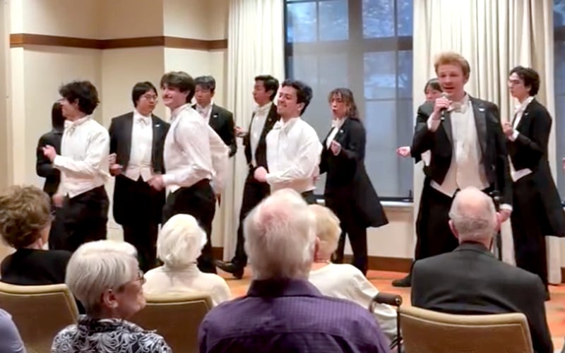 A group of young men in formal attire sing and perform in front of an audience of elderly people seated in a room with large windows.