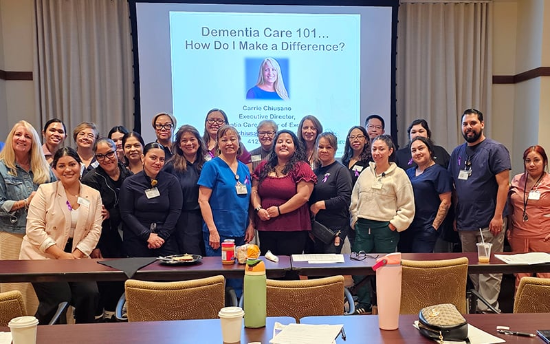 A group of people pose together in a conference room in front of a screen displaying “Dementia Care 101 – How Do I Make a Difference?” Tables with drinks and notepads are in the foreground.