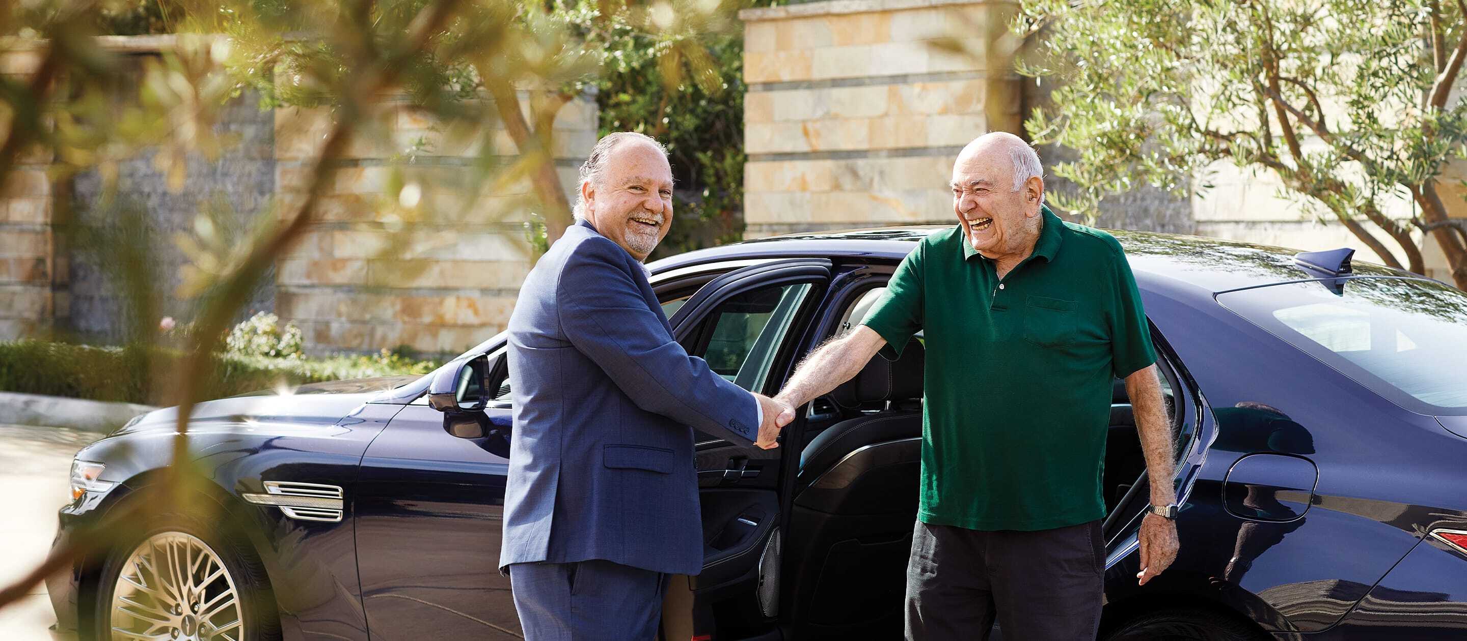 Two older men smile and shake hands next to an open car door, one wearing a suit and the other in a green polo shirt, with trees and a stone building in the background.