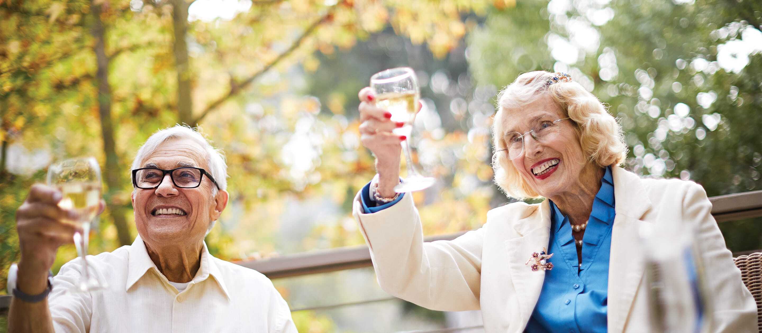 Two smiling older adults sit outdoors, raising glasses of white wine in a toast. They are dressed smartly and appear happy, with autumn-colored trees in the background.