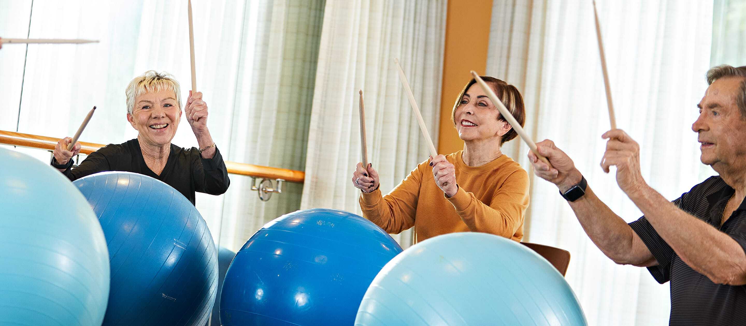 Three older adults sitting and smiling while holding drumsticks, using large blue exercise balls as drums in a bright, airy room with curtains and wooden accents.