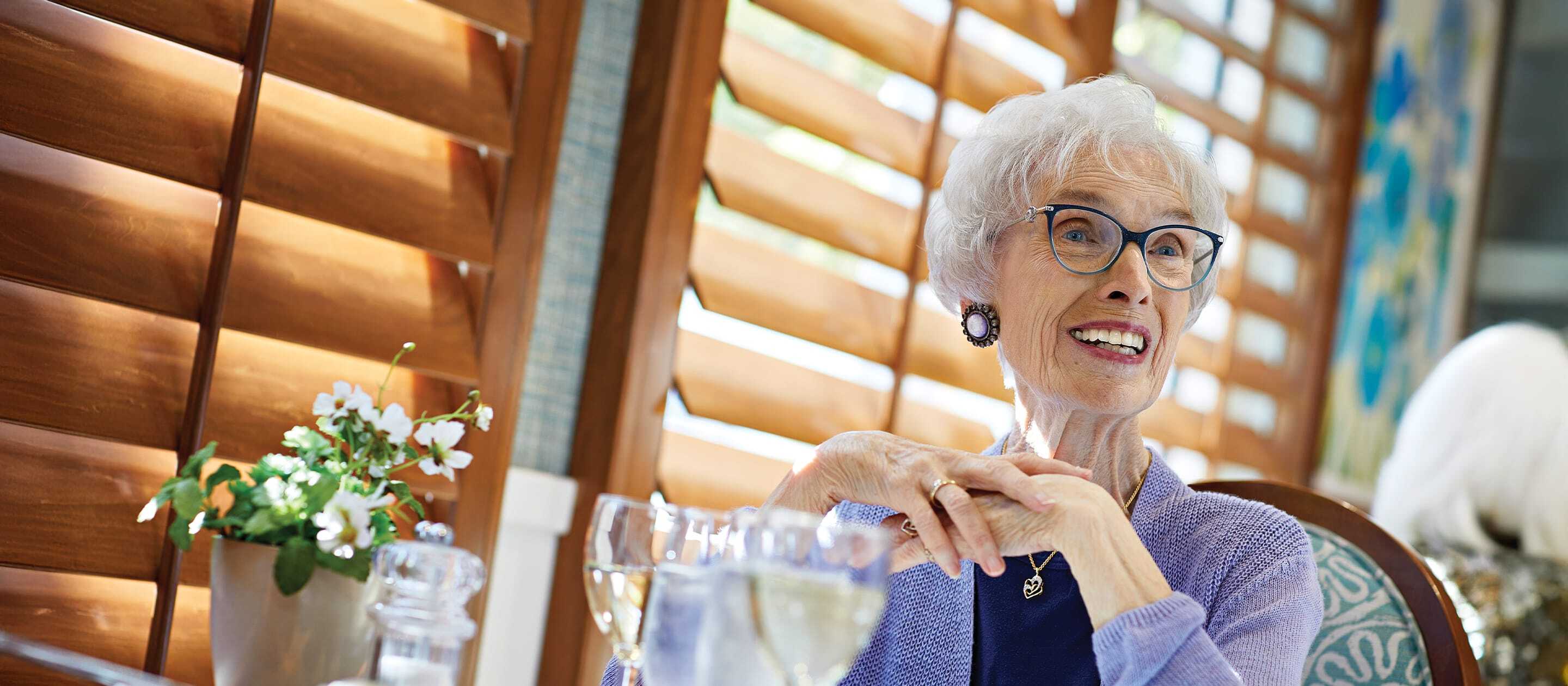 An elderly woman with white hair and glasses smiles while sitting at a table with a glass of water; sunlight streams through wooden blinds behind her, and a small plant is nearby.