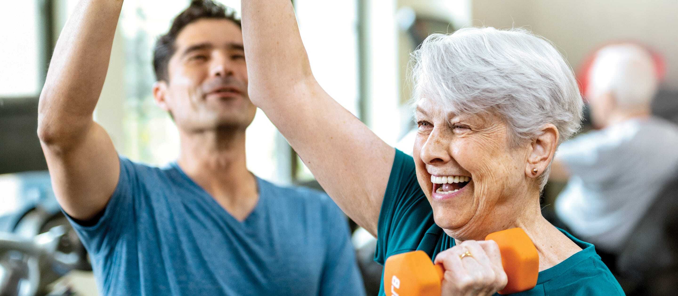 An older woman with short gray hair smiles while lifting orange dumbbells, assisted by a man in a blue shirt at a gym. Both appear happy and engaged in the fitness activity.