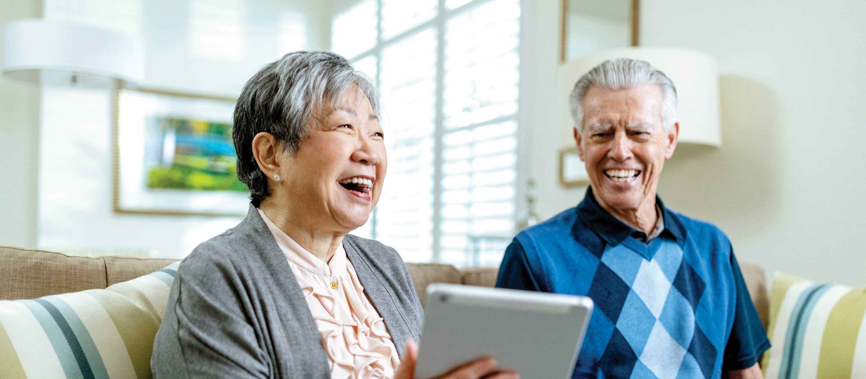 Two smiling older adults sit on a couch; the woman holds a tablet while the man looks on. Bright light comes through a window behind them, creating a cheerful, cozy atmosphere.