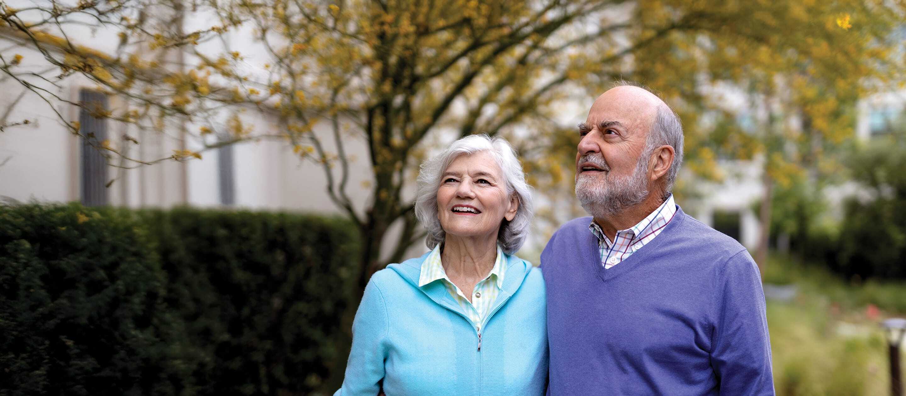 An older couple stands outdoors, smiling and looking up. The woman wears a light blue sweater over a collared shirt, and the man wears a purple sweater over a shirt. Trees with yellow leaves and greenery are in the background.