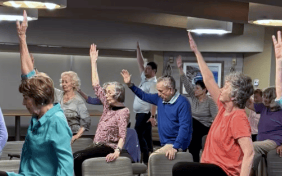 A group of older adults sit in chairs and participate in a seated exercise class, raising one arm in the air. The room is well-lit with a relaxed atmosphere.