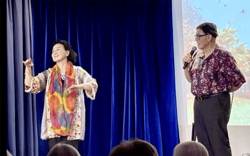 Two people stand on a stage with a blue curtain; one speaks into a microphone while the other gestures expressively with their hands, wearing a colorful scarf and patterned top.
