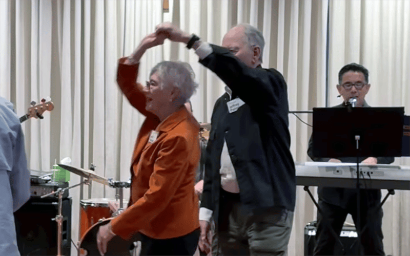 Two older adults dance together, smiling, while a musician plays keyboard and sings in the background. Drums and other instruments are visible behind them. The scene appears festive and joyful.