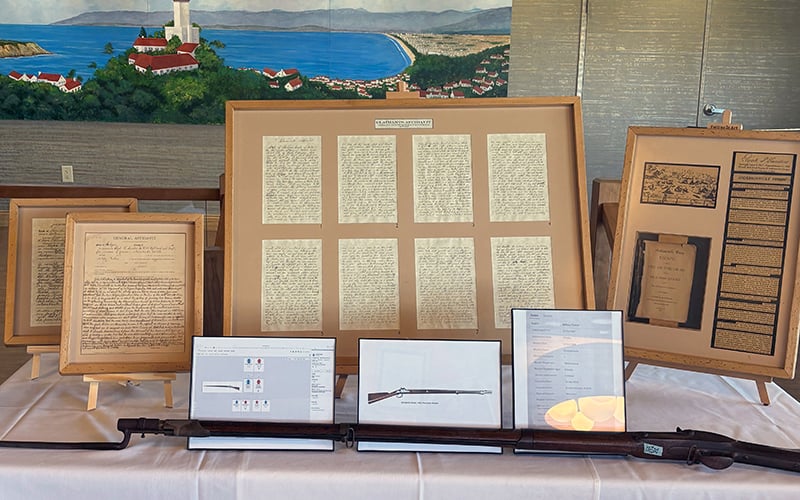 A display table with historical documents in frames, illustrations of rifles, museum-style labels, and a rifle laid out in front. Behind the display is a mural of a coastal town with a lighthouse and ocean.
