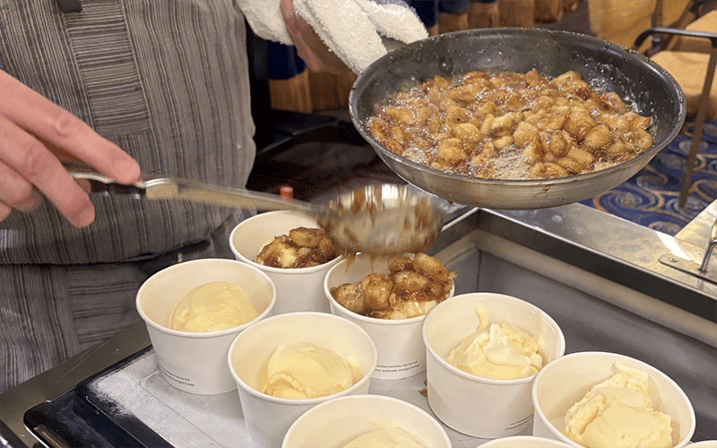 A person spoons caramelized bananas from a skillet onto cups of vanilla ice cream, preparing banana foster desserts. The cups are arranged in rows on a tray.