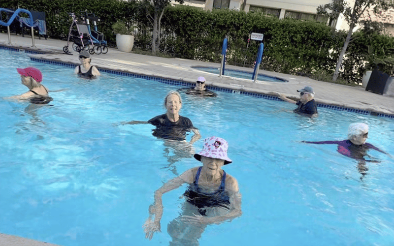 Group of older adults participating in an aqua aerobics class in an outdoor swimming pool. Most are wearing hats and swimsuits, and some are smiling while exercising in the water. A walker is visible by the poolside.