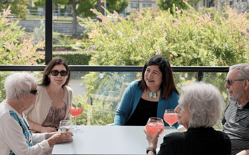 Five adults sit together at an outdoor table, enjoying drinks. Greenery surrounds them and sunlight filters in. The group is engaged in conversation, with three women and two men smiling and relaxed.