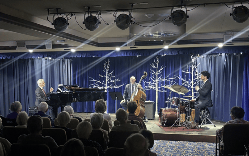 A jazz trio performs on stage with a pianist, upright bassist, and drummer. The audience is seated in front of them. The stage features blue curtains, white decorative trees, and dramatic overhead lighting.
