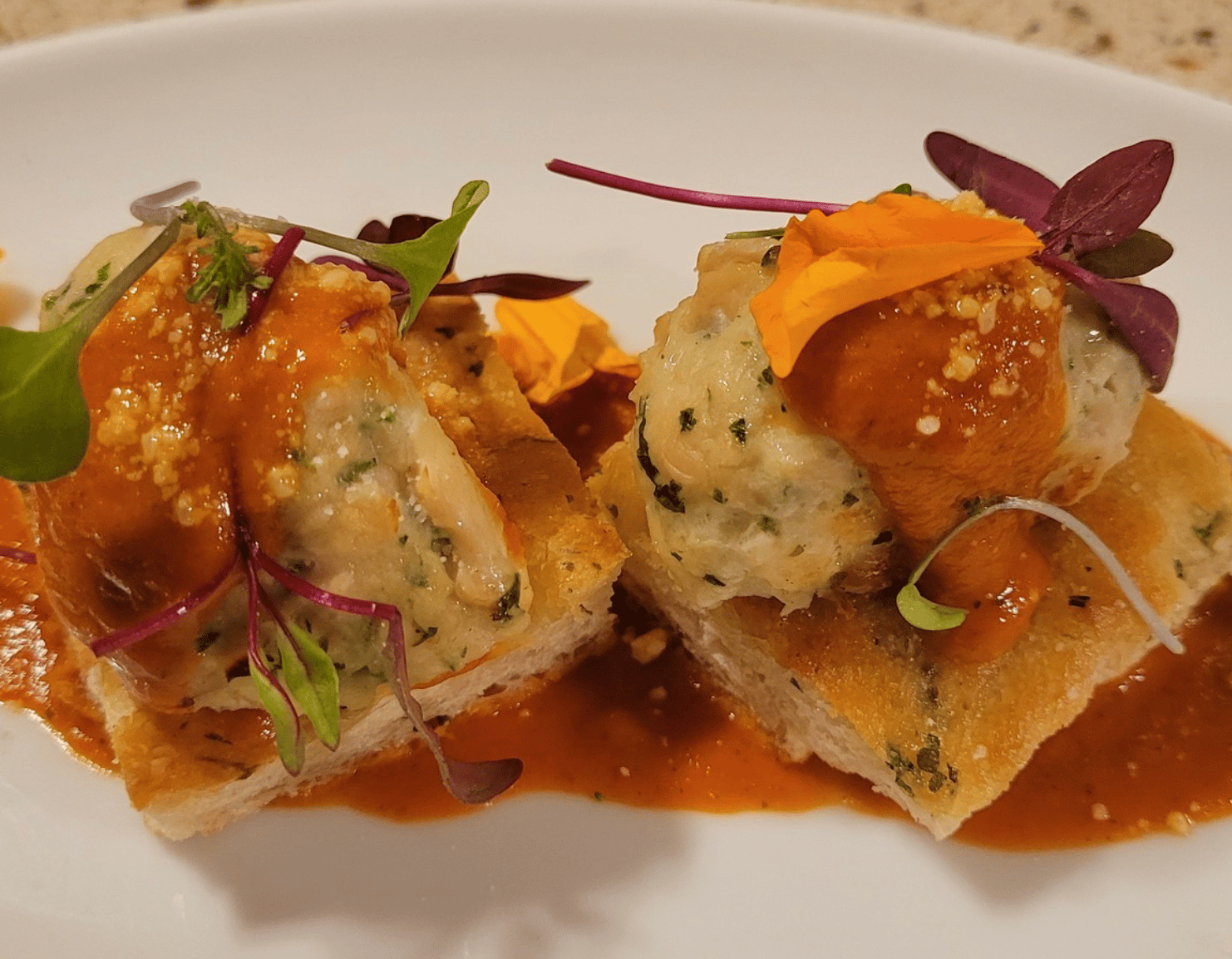 Two pieces of herbed bread topped with savory dumplings, garnished with microgreens and orange flower petals, and drizzled with a rich red sauce, served on a white plate.