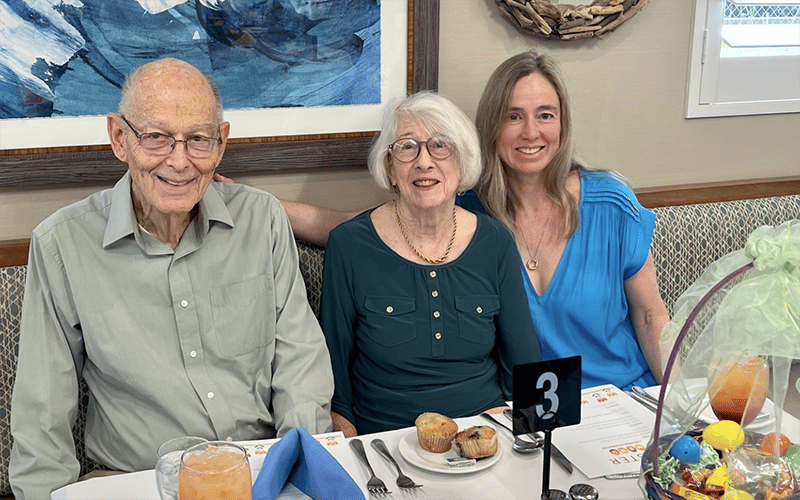 Three people sit together at a dining table, smiling at the camera. The table has drinks, muffins, and an Easter basket. There is a menu and a table number 3 sign in front of them. Art hangs on the wall behind them.