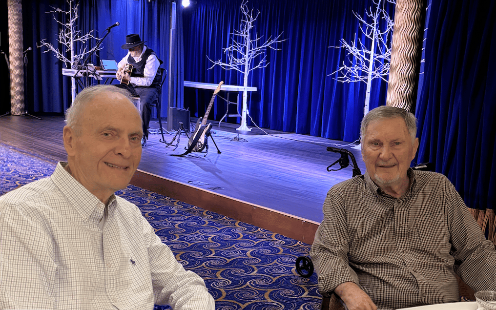 Two older men sit and smile at a table in front of a stage with blue curtains. On stage, a musician in a hat plays guitar near bare decorative trees and empty chairs. The setting has patterned carpeting.