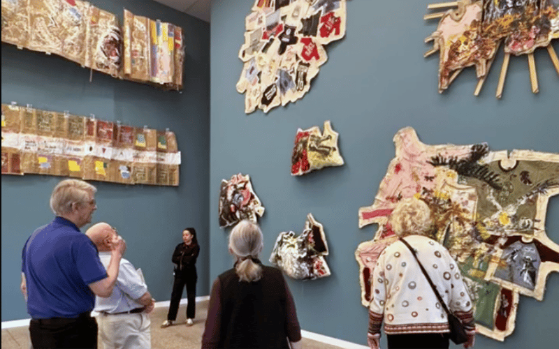 A group of people view colorful, textured abstract art pieces mounted on large blue walls in a gallery. The artworks vary in shape and size, with a gallery guide standing nearby.