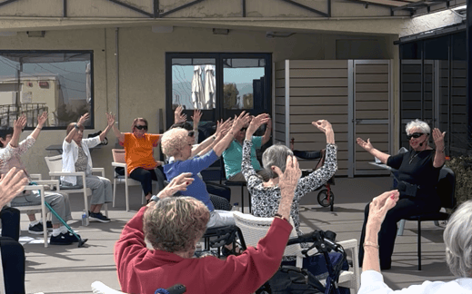 A group of older adults participate in a seated exercise class outdoors, raising their arms while following an instructor at the front. The setting appears to be a patio at a care facility or community center.