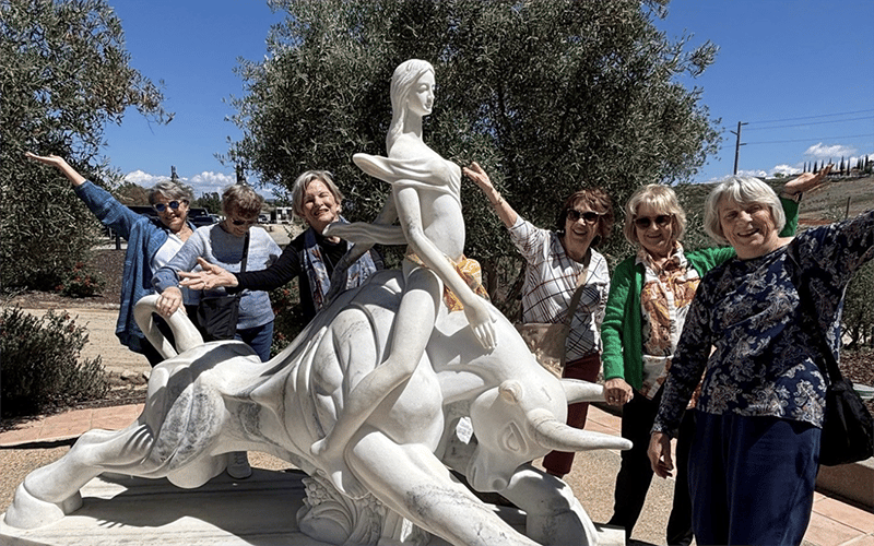 Six smiling women stand outdoors around a large white sculpture of a woman riding a bull, posing with their arms raised playfully. Trees and blue sky are visible in the background.