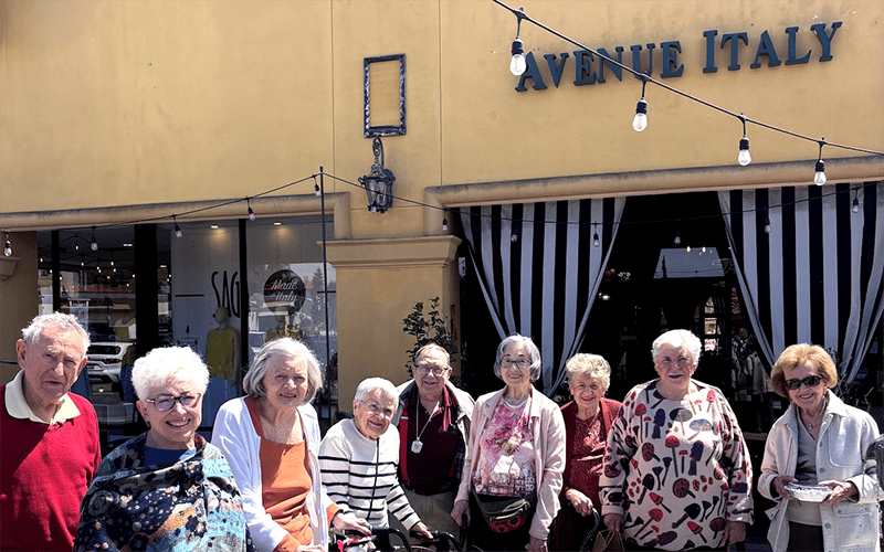 A group of nine smiling older adults stands together outside a yellow building with a sign that reads Avenue Italy and striped black-and-white curtains at the entrance. String lights hang above them.