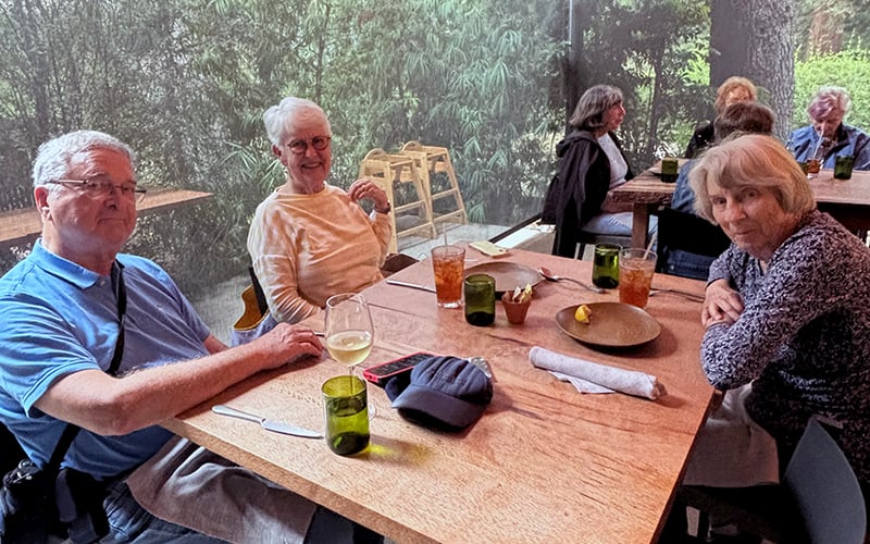 Four older adults sit around a wooden table at a restaurant, smiling and enjoying drinks. Empty plates and glasses are on the table. A lush green view is visible through large windows in the background.