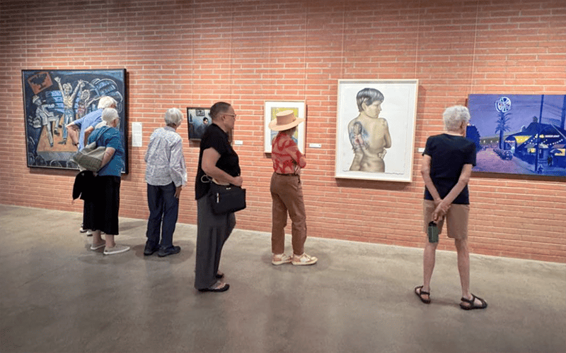A group of six people stands in a gallery, observing various paintings hung on a brick wall. The visitors are casually dressed and appear to be engaged with the diverse artwork on display.