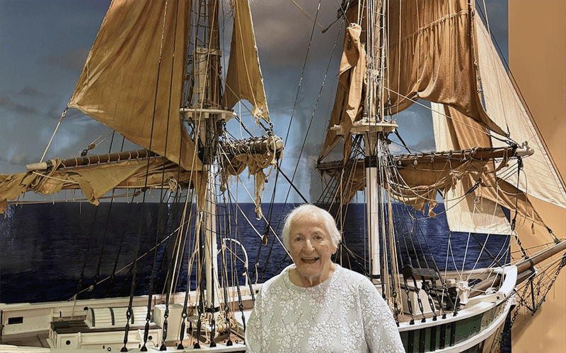 An elderly woman in a white blouse stands smiling in front of a large model of a historic sailing ship, with the ocean and sky depicted in the background.