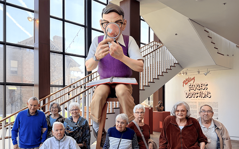 A group of elderly people pose in front of a large sculpture of a man with glasses reading a book with a magnifying glass inside a bright, modern museum with big windows and a staircase.