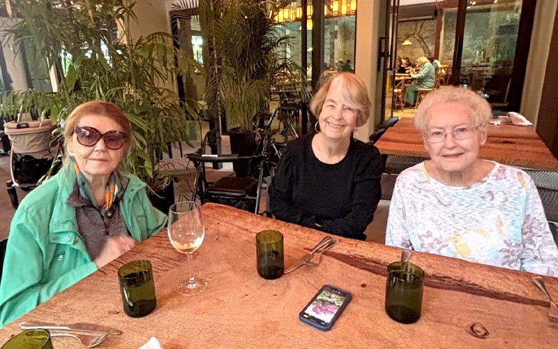 Three older women sit at a restaurant table with empty glasses and a phone, smiling at the camera. They are casually dressed, and plants and other diners are visible in the background.