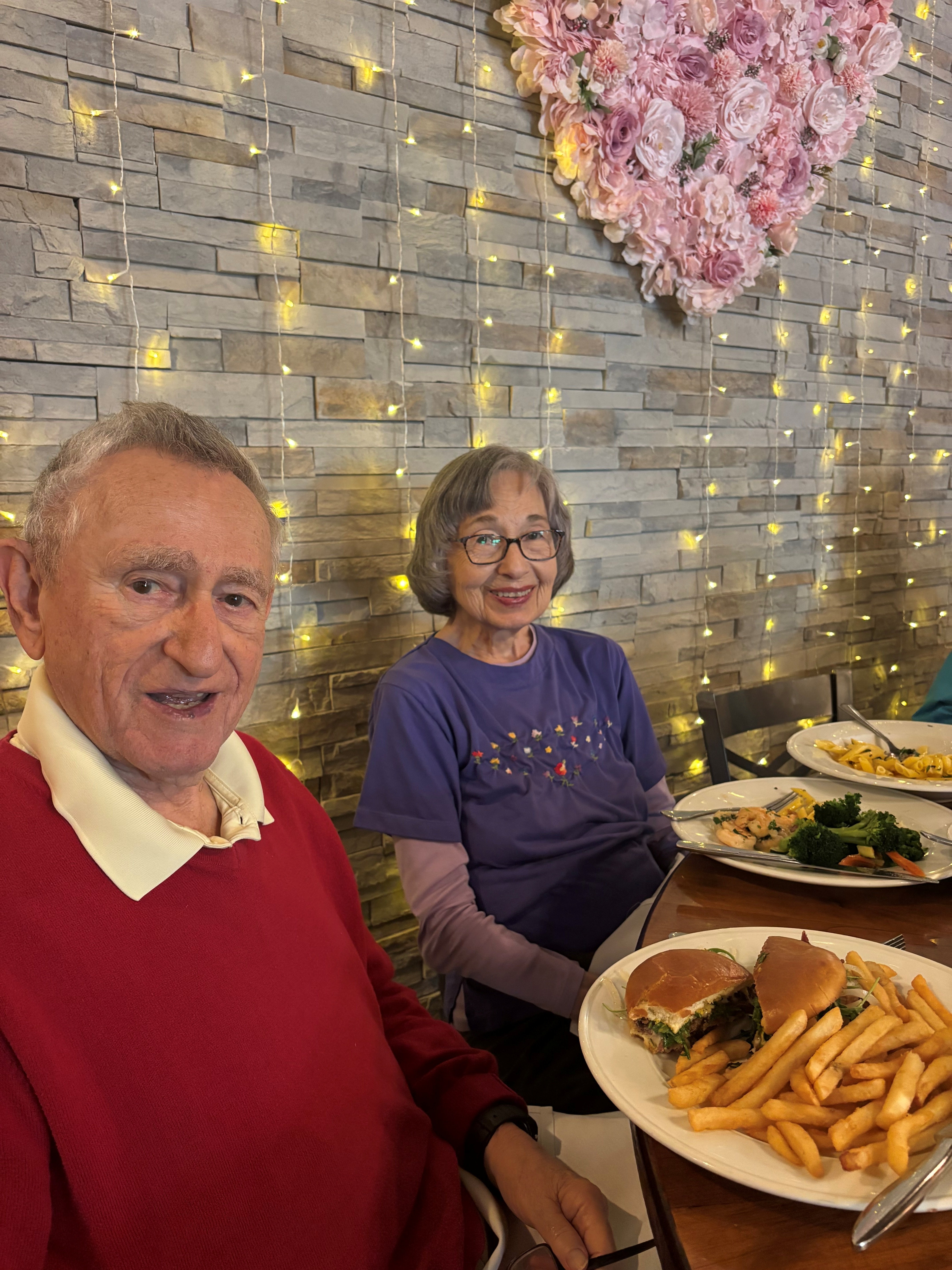 An elderly man and woman sit at a restaurant table with plates of food, including burgers and fries. Behind them is a stone wall decorated with string lights and a large heart made of pink flowers.