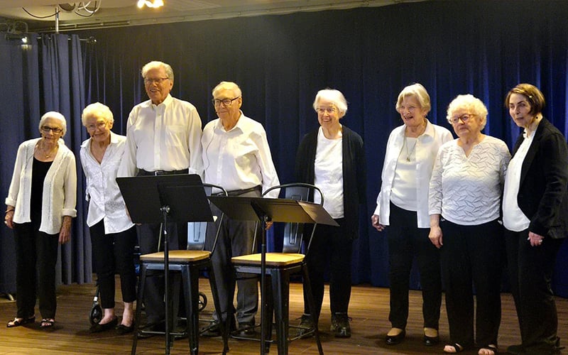 A group of eight older adults, dressed in black and white clothing, stand side by side on a wooden stage with dark blue curtains behind them. Two empty chairs and music stands are positioned in front of them.