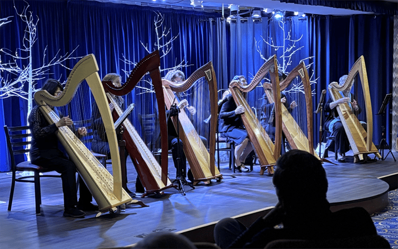 A group of six harpists perform on stage under blue lighting, each seated behind a harp. Bare white tree branches decorate the background, and audience members watch from the foreground.