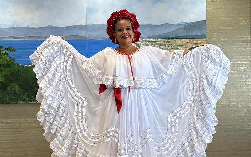 A woman smiles while wearing a traditional white folkloric dress with lace details and a red ribbon, along with a matching red headpiece. She holds out the sides of her dress, displaying its wide, flowing skirt. A scenic mural is behind her.