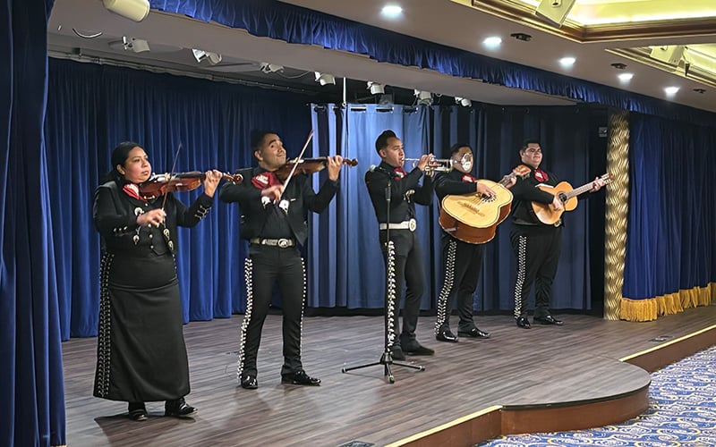 Five mariachi musicians perform on stage in traditional black outfits with red ties, playing violin, trumpet, guitarrón, and guitar, with blue curtains and gold trim in the background.