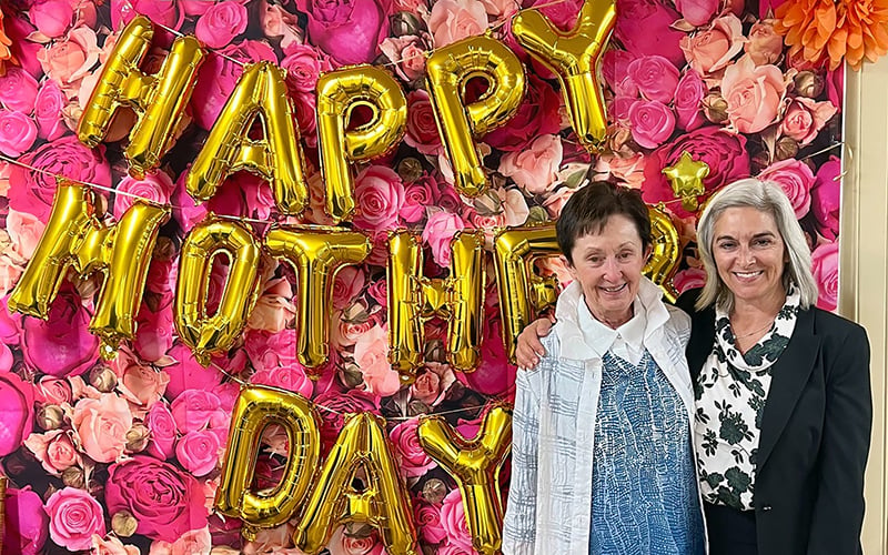 Two women stand smiling in front of a wall decorated with pink roses and large gold balloons spelling Happy Mothers Day. One woman is elderly, the other has gray hair and wears a black blazer.
