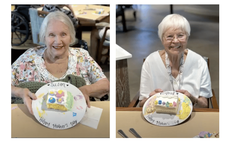 Two elderly women, each smiling and holding a decorated plate with a small cake and Happy Mothers Day written on it. One plate says Susan and the other says Carol.