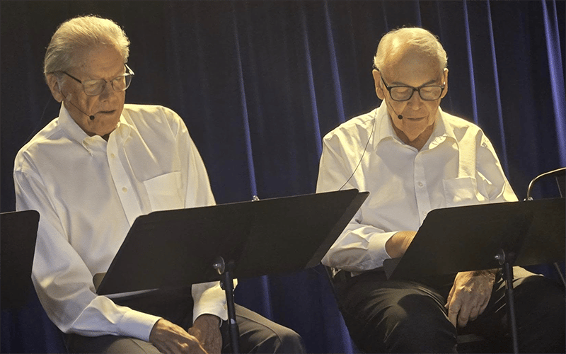 Two elderly men wearing glasses and white shirts sit side by side on stage, reading from black music stands, with a dark blue curtain in the background. Both have microphones on their faces.