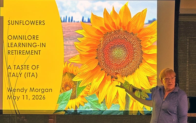 A woman stands in front of a projected slide showing a sunflower and text about an Omnilore learning-in-retirement event titled A Taste of Italy by Wendy Morgan, scheduled for May 11, 2026.